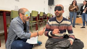 En primer plano, aparecen dos profesores del IES Palomeras-Vallecas intentando escribir mientras están con los ojos cubiertos por un antifaz. Detrás, aparecen la profesora y la alumna con discapacidad visual de la Universidad Rey Juan Carlos que dirigen la actividad.