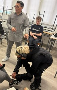 Un alumno del IES Palomeras-Vallecas con un traje de esfuerzo que le permite experimentar lo que siente una persona con una discapacidad que le afecta a la fuerza intenta atarle los cordones de una zapatilla a una compañera. El alumno lleva puestas también unas gafas que simulan cómo ve una persona con una determinada discapacidad visual y unos cascos que no le permiten oír bien. Es observado por un profesor de la Universidad Rey Juan Carlos, que dirige la actividad, y por un alumno de la misma universidad con discapacidad motriz.