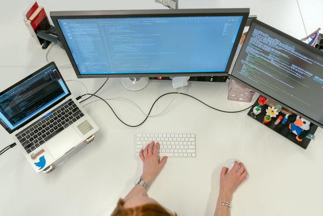 Un ordenador con dos pantallas sobre una mesa y unas manos escribiendo en el teclado