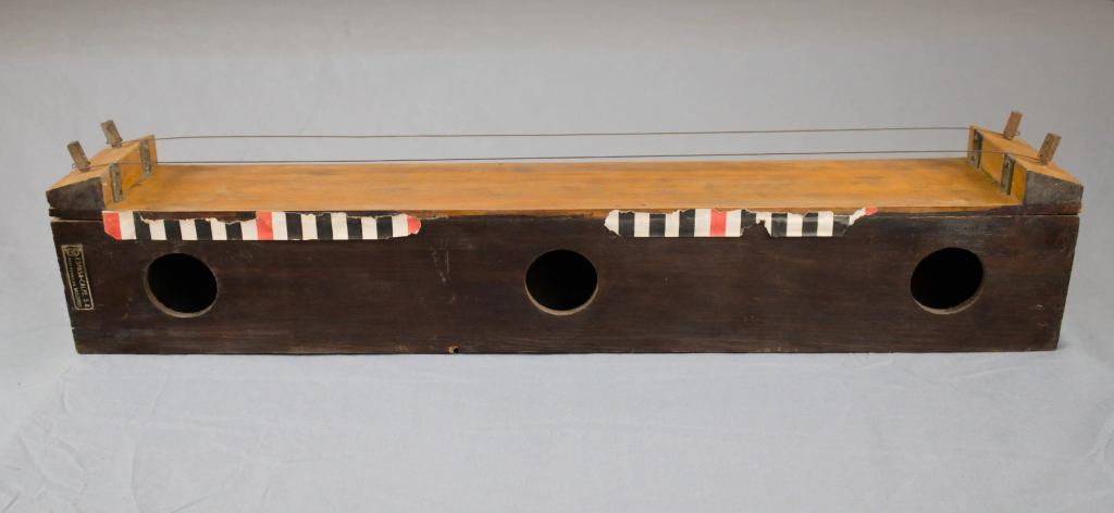 Fotografía de instrumento con forma de caja de madera. Se ve chapa con la marca del fabricante a la izquierda. Cuerdas en la parte superior y aperturas circulares de resonancia en el frontal.